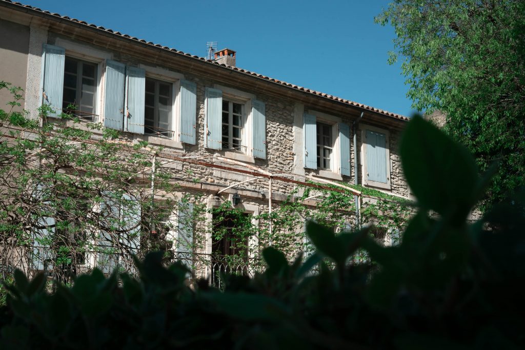 A historical building with stone walls and light blue shutters, partially obscured by lush greenery in the foreground, creating a serene and picturesque setting.