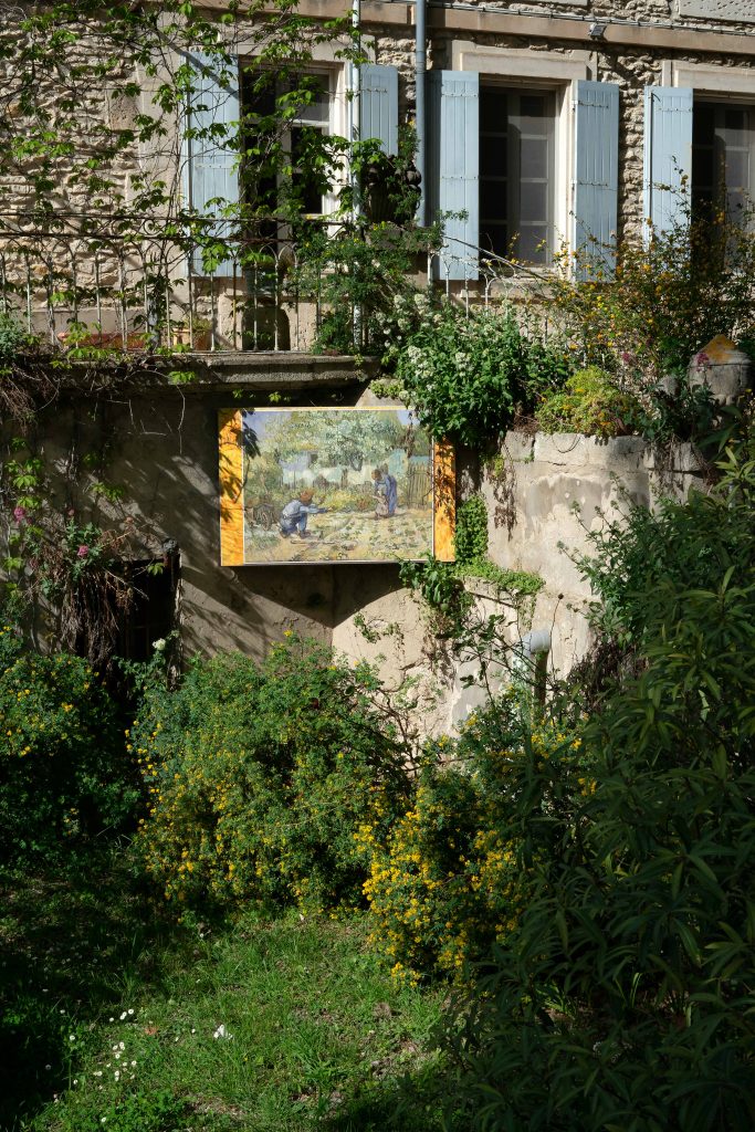 A picturesque scene of a lush garden with flowering shrubs, with a large framed painting depicting a rural landscape hanging on an old, weathered stone wall beneath blue shuttered windows at the Monastery Saint-Paul de Mausole.