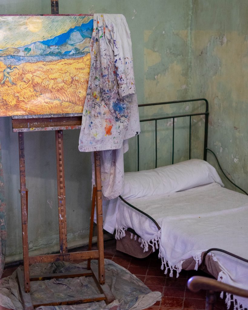 A painter's easel displaying a vibrant landscape painting, with a paint-stained smock draped over it, set against a backdrop of a rustic room with peeling green paint and a single, simple bed covered by a white sheet.