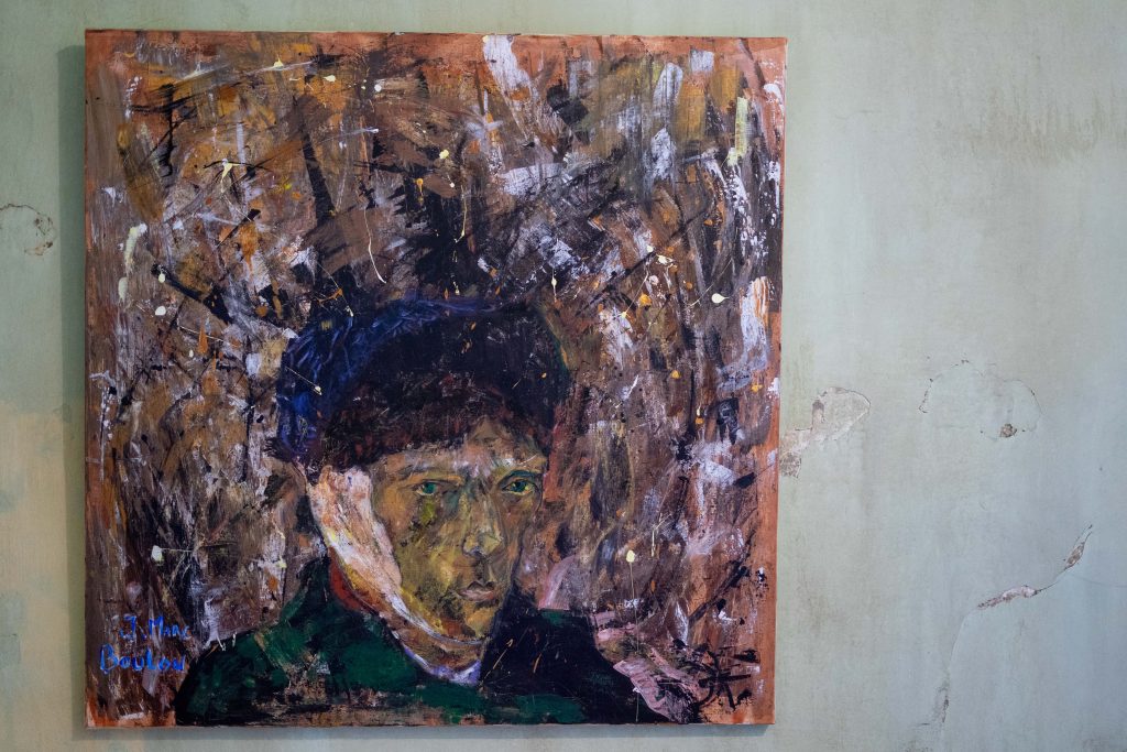 A textured, expressionistic painting of a man's face, prominently displayed on a green wall with visible signs of wear and peeling. The artwork features bold, chaotic brush strokes in earthy tones surrounding the more detailed, somber face.