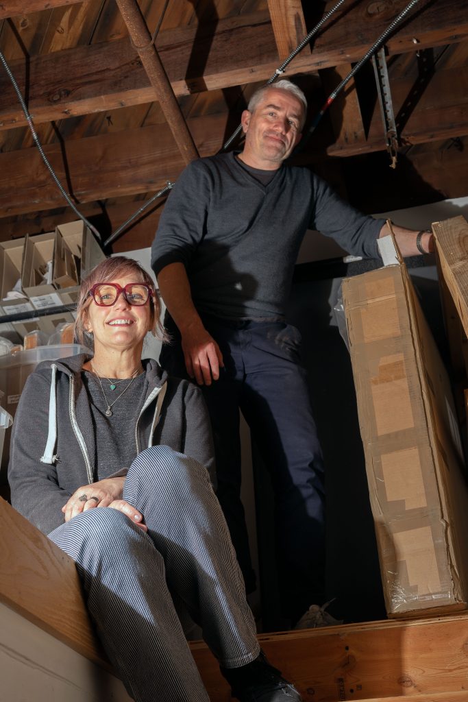 A man and a woman in an attic filled with stored items. The woman is seated comfortably on a wooden ledge, smiling at the camera, while the man stands behind her, leaning on a large cardboard box, also smiling. Both are surrounded by wooden beams and storage boxes.