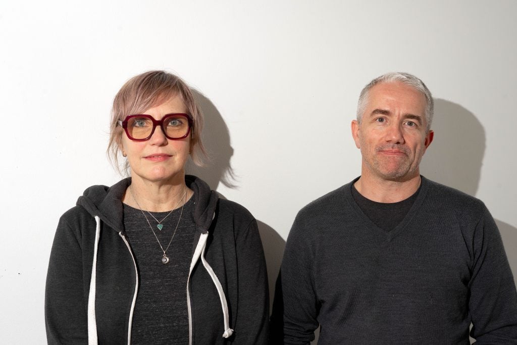 A portrait of a man and a woman standing side by side against a plain white wall. The woman on the left wears glasses and a dark hoodie, while the man on the right sports a gray sweater. Both are looking directly at the camera, casting subtle shadows on the wall behind them.