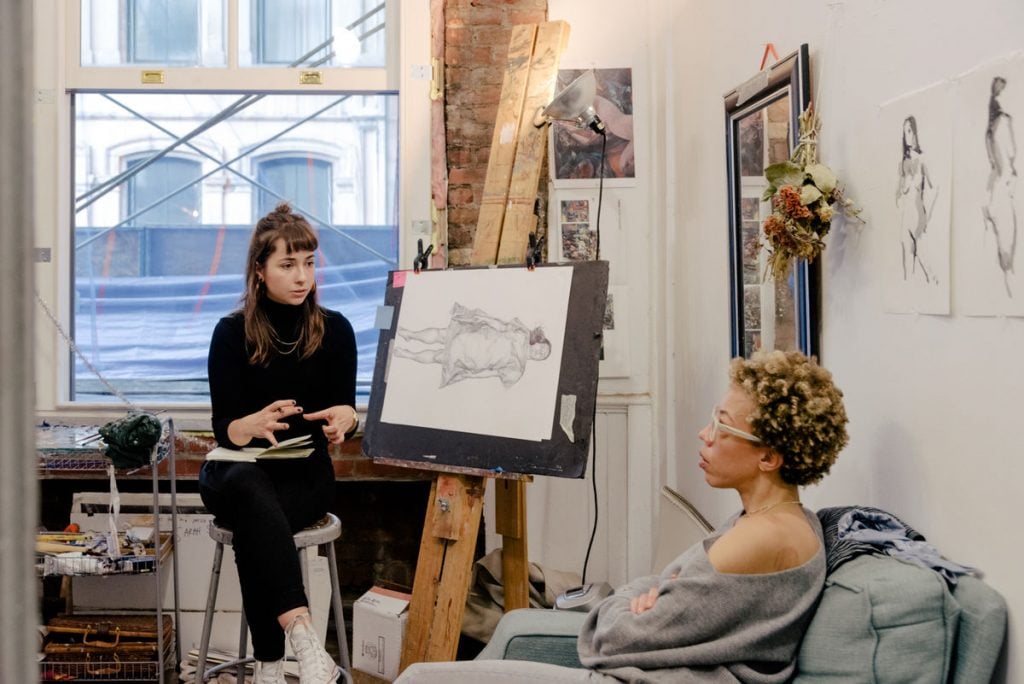 A woman sitting in a studio by an easel speaking to another woman sitting on a nearby sofa