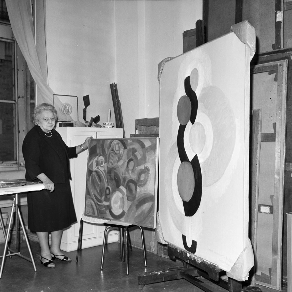 An elderly lady standing in a studio alongside two of her paintings