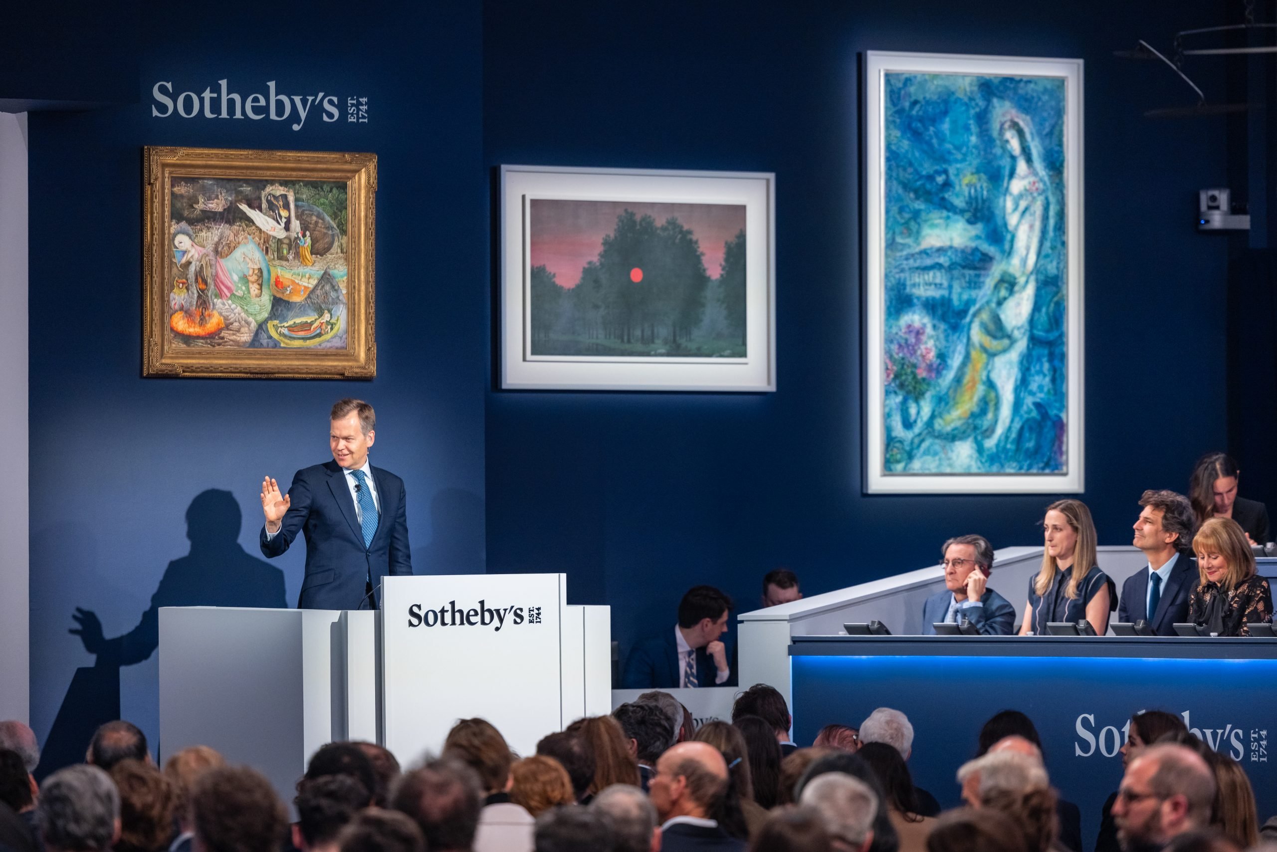 The sale room at Sotheby's Modern Evening Art Sale with a man standing at a podium that reads "Sotheby's," three paintings hanging behind him, and a group of phone bidders to his left. An audience sits in the foreground