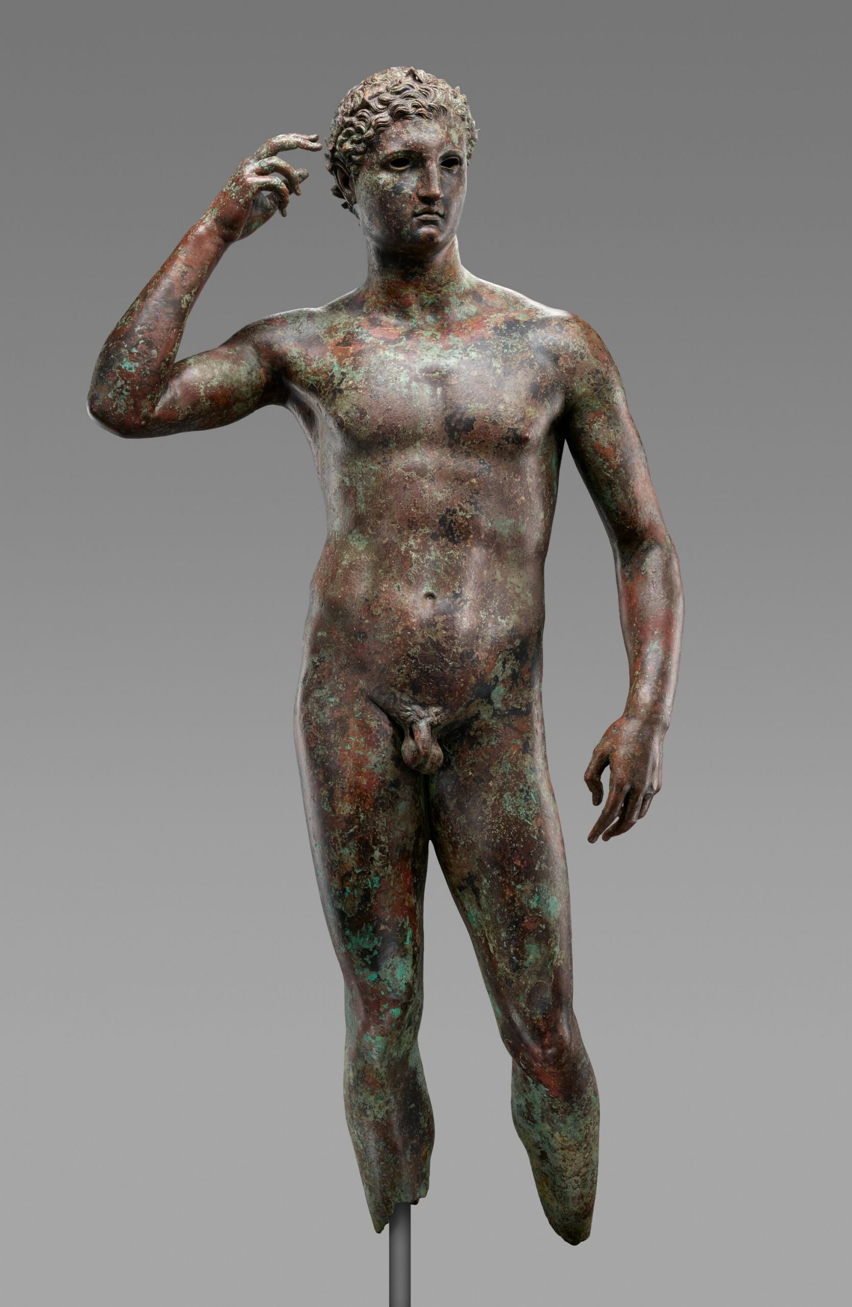 Ancient bronze statue of a young male athlete, known as the "Victorious Youth." The figure stands in a relaxed pose, raising his right hand to adjust a wreath on his head, showing intricate details and a naturalistic body form, with a patina that hints at its age and historical significance.