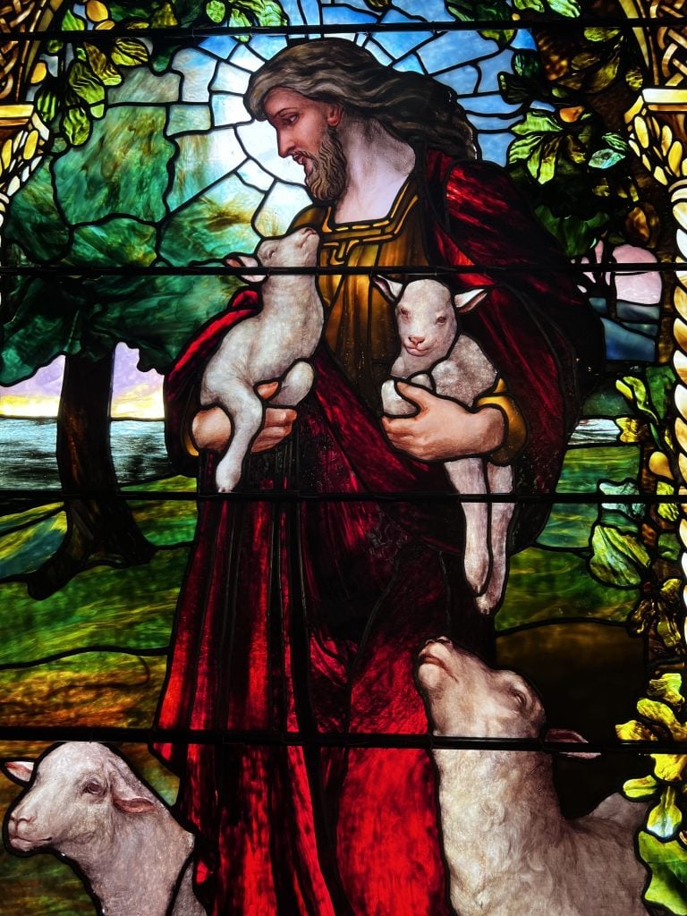 A stained glass window showing Christ in red robes holding two lambs, with two sheep at his feet