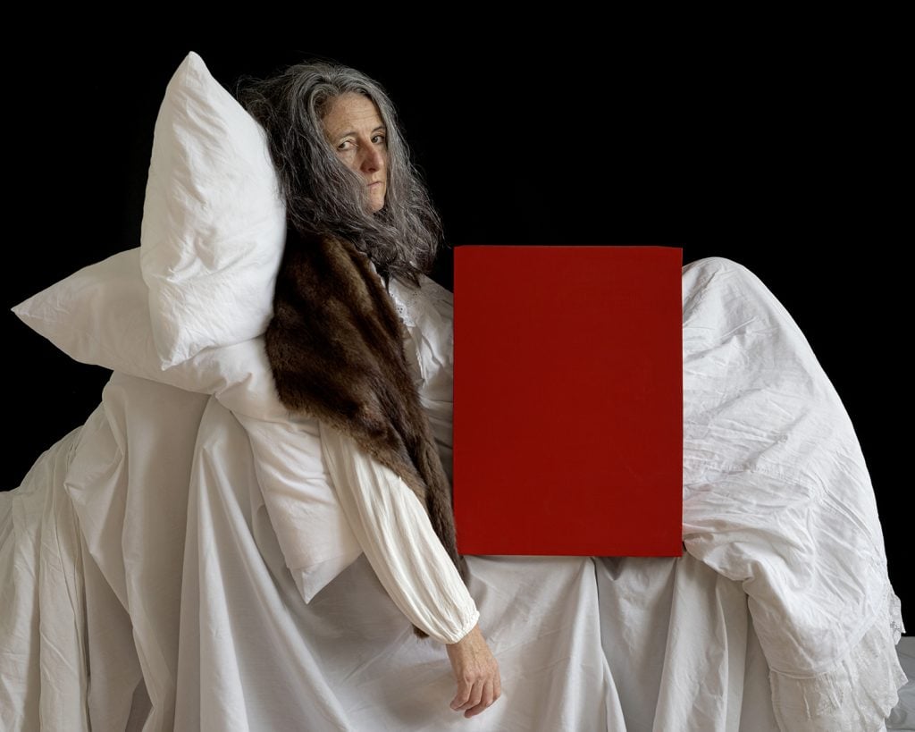 A self-portrait of Trish Morrissey in character in a shite nightgown on a bed in white linens against a black background with a large red box over her middle section.