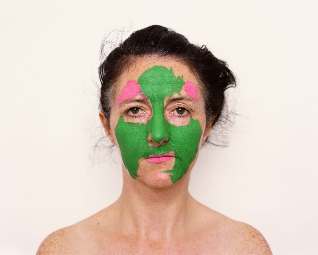 A headshot self-portrait of artist Trish Morrissey in front of an off-white background and bare shoulders with green and pink paint smeared on her face.