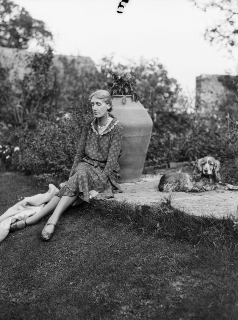 black and white photograph of Virginia Woolf sitting on a grassy bank in front of a large urn