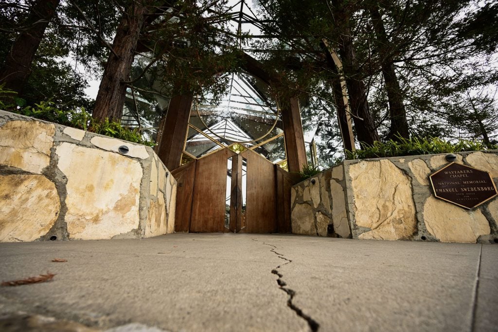 An image of the entrance to Wayfarers Chapel, also known as the National Memorial to Emanuel Swedenborg. The perspective is from the ground, looking up at the chapel’s wooden doors, framed by stone walls and tall trees. The entrance features a plaque with the chapel's name and a visible crack in the concrete walkway leading up to the doors.