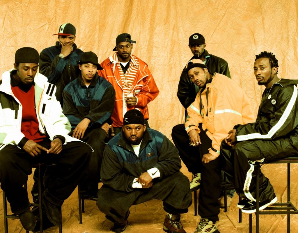 Eight men posing against a yellow backdrop.