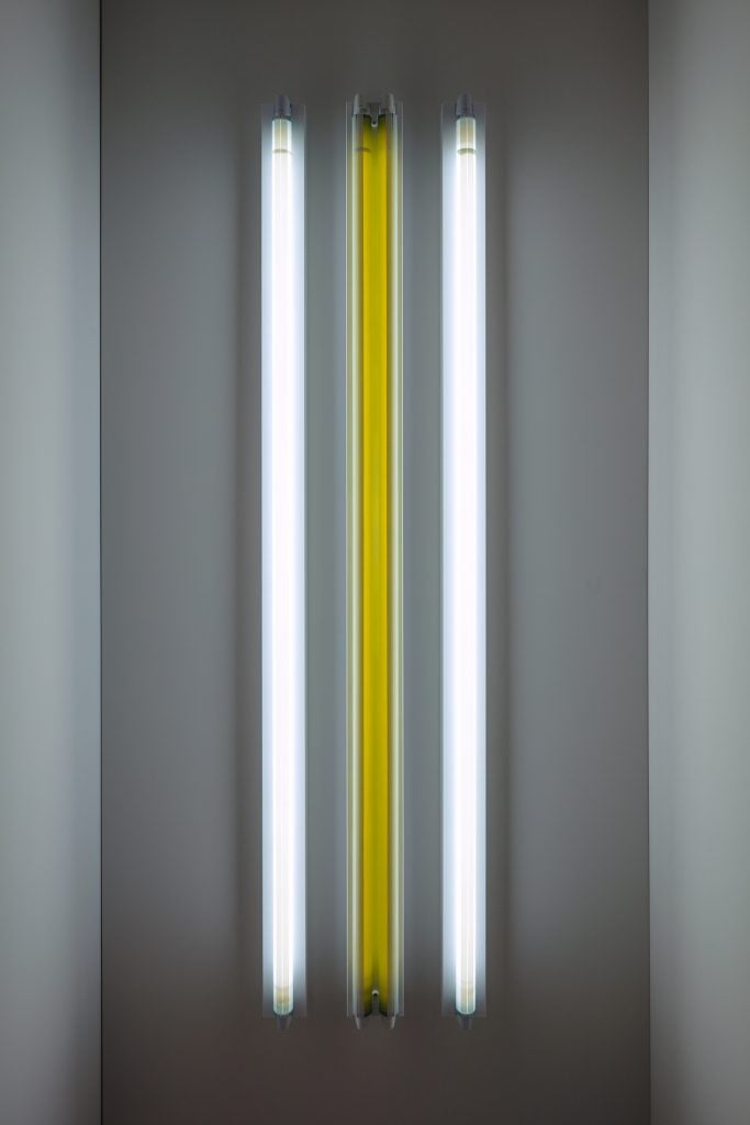 The image shows an art installation consisting of three vertical fluorescent light tubes mounted on a wall. The arrangement features two white light tubes on the outer sides and a yellow light tube in the center. The lights create a minimalist and modern visual effect, highlighting the contrast between the colors and the simplicity of the design. The installation is likely part of a contemporary art exhibit, focusing on light and color as primary elements.