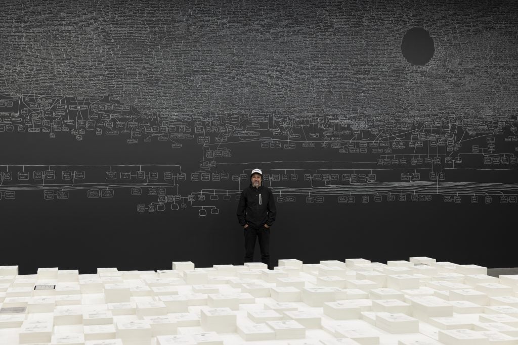 A man standing in front of a white installation against the backdrop of.black board with images drawn by chalk