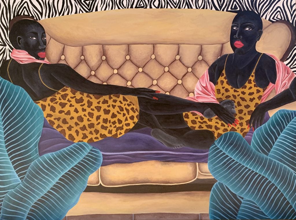 two black women dressed in patterned dresses reclining on a sofa look directly at the viewer