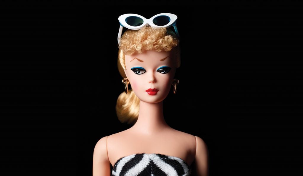 Image of the original Barbie 1959 in strapless black and white stripy bathing suit and white framed sunglasses, against a black backdrop