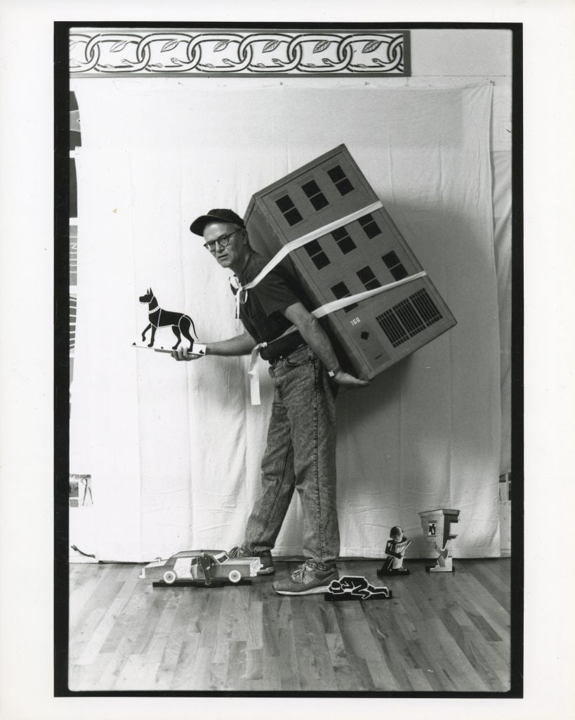 A black-and-white photo features an elderly man wearing glasses and a cap, performing in an art piece. He is strapped with a large building model on his back and holding a cut-out figure of a dog. On the floor are several other cut-out figures, including a car and a person. The scene is set against a plain backdrop with a decorative border at the top, creating a theatrical and whimsical atmosphere.