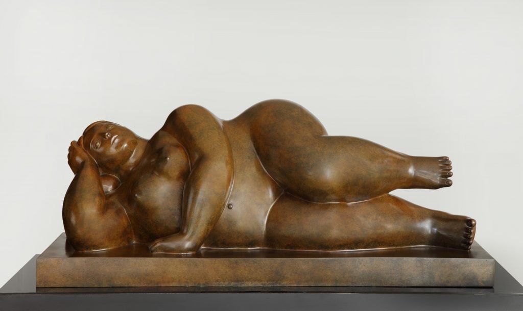 A bronze sculpture by Fernando Botero titled "Reclining Woman," created in 2007, depicting a voluptuous woman lying on her side with her head resting on her hand, characteristic of Botero's signature style of exaggerated and rounded forms.