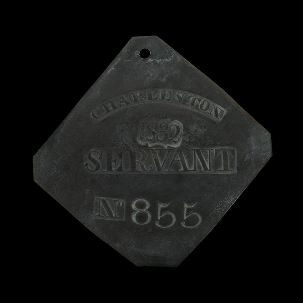 The image shows a historical slave badge from Charleston, South Carolina, dated 1832. The badge is diamond-shaped with the following inscriptions: "CHARLESTON" at the top. The year "1832" below Charleston. "SERVANT" indicating the occupation. The number "No 855" at the bottom, serving as a unique identifier. These badges were used to identify enslaved individuals who were hired out for work by their owners, regulating and controlling the system of urban slavery. The badge features a hole at the top, likely for attaching to the individual as a form of identification. This specific badge provides insight into the methods used for managing and documenting the labor of enslaved people during that period. 2/2