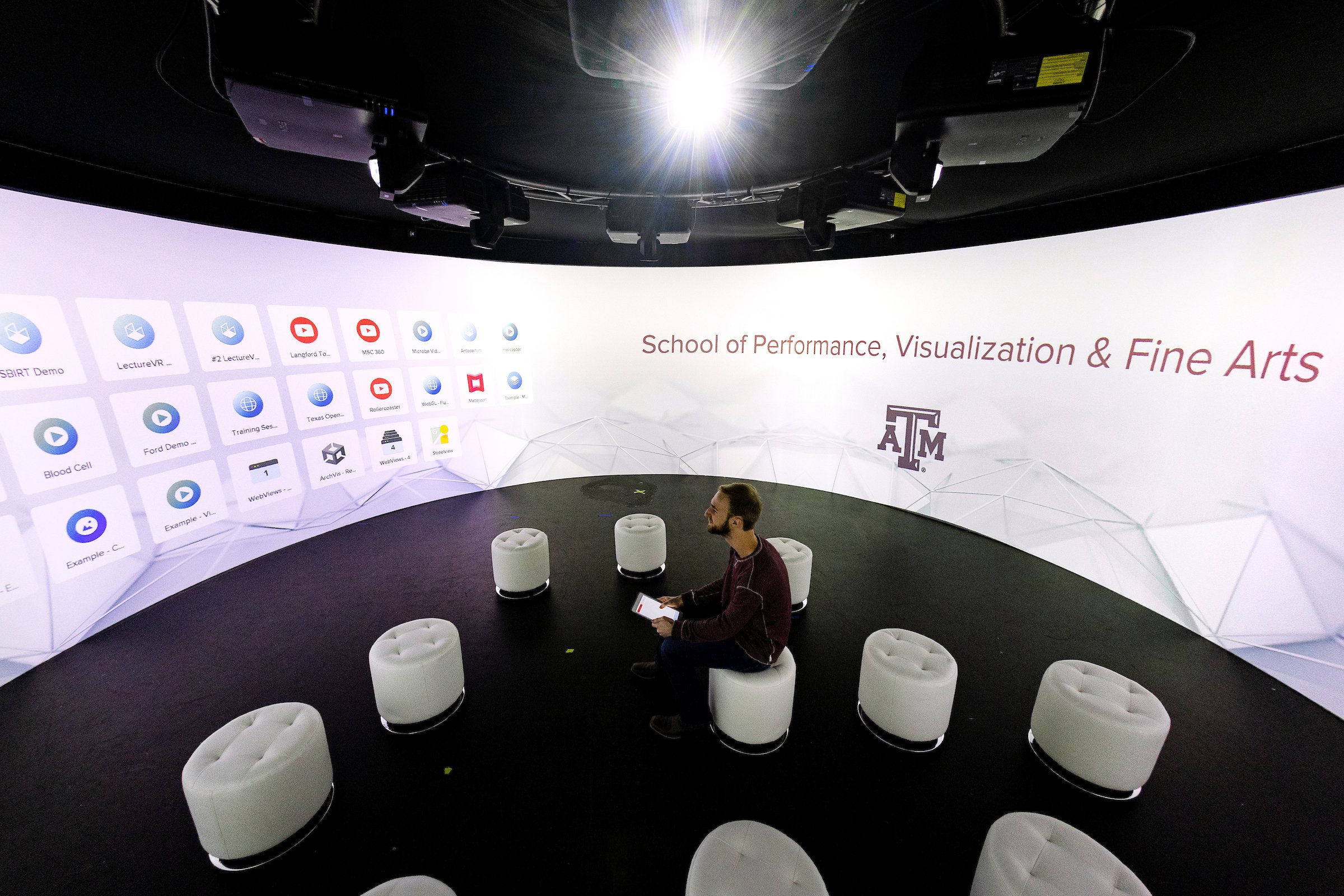 A person sits in a high-tech immersive environment, surrounded by a curved screen displaying various icons and applications. The space is part of the School of Performance, Visualization & Fine Arts at Texas A&M University. Several white, cylindrical stools are arranged in the room, emphasizing its modern and innovative design.