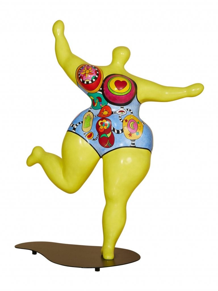 A vibrant sculpture by Niki de Saint Phalle titled "Dawn Jaune," created in 1995. The piece features a joyful, exaggerated female figure in bright yellow, adorned with colorful, whimsical decorations and symbols on her torso, typical of de Saint Phalle's playful and bold style.