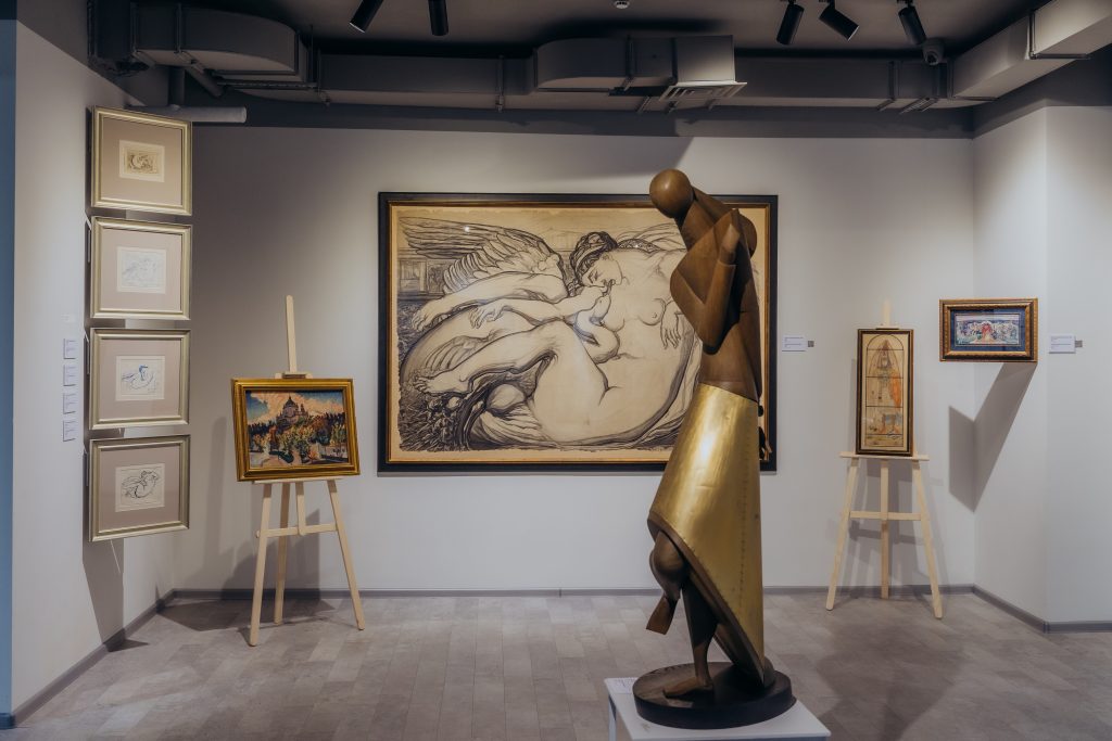 A fragment of an art exhibition, showing a variety of artworks displayed on walls and easels. The foreground features an abstract wooden sculpture. In the background, there are several framed paintings, including a colorful abstract portrait and a depiction of a mother with a child. The exhibition space is well-lit, highlighting the details and colors of the artworks.