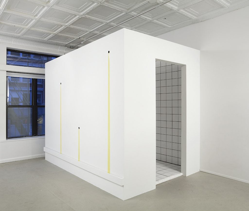 A small white room stands in another white room. Yellow liquid drips down its side in three places.