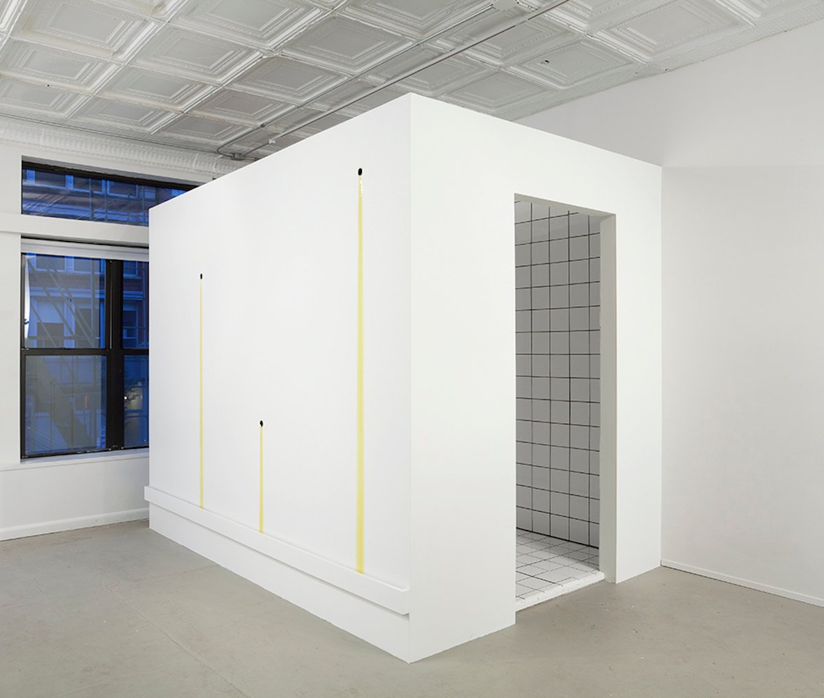 A small white room stands in another white room. Yellow liquid drips down its side in three places.