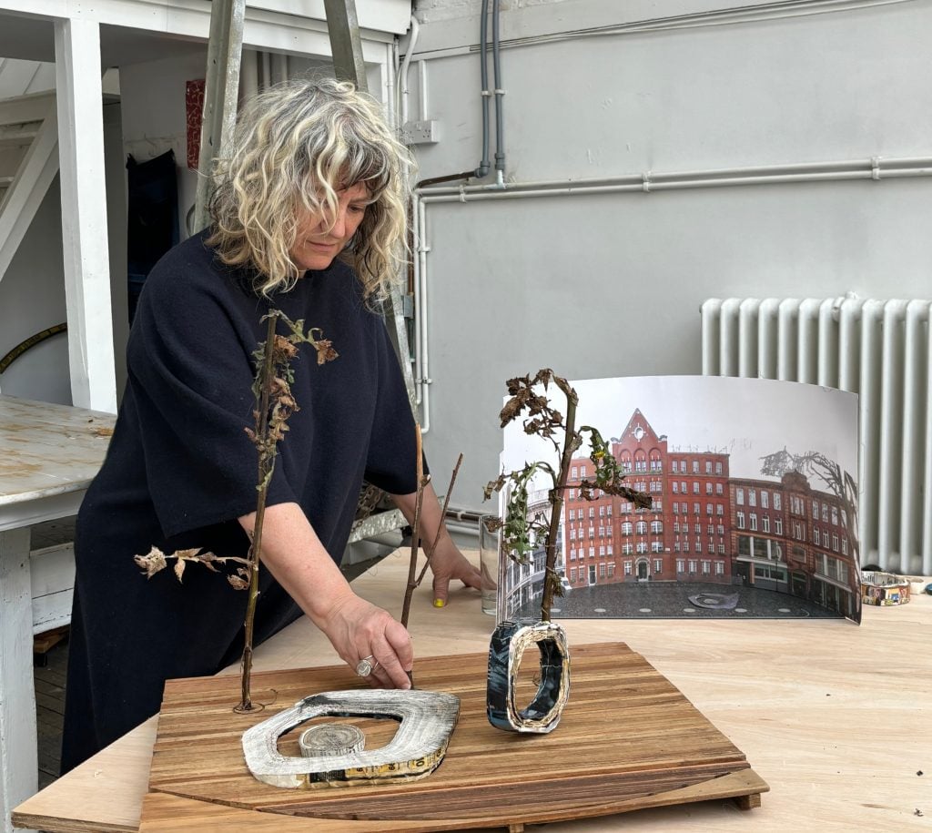 a woman stands in front of a wooden board with a miniature model of a memorial that looks like parts taken from the cross section of a tree trunk, some twigs with fronds are positioned around in the manner of trees