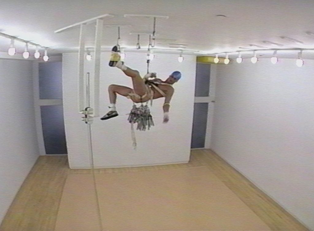 A partially nude man hangs from the ceiling with rope