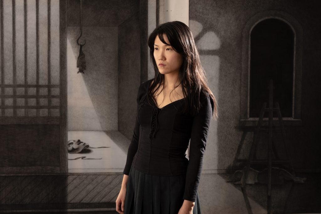 a woman with long dark hair in a black dress stares out into the distance, as shadowed filled artwork is visible behind her.