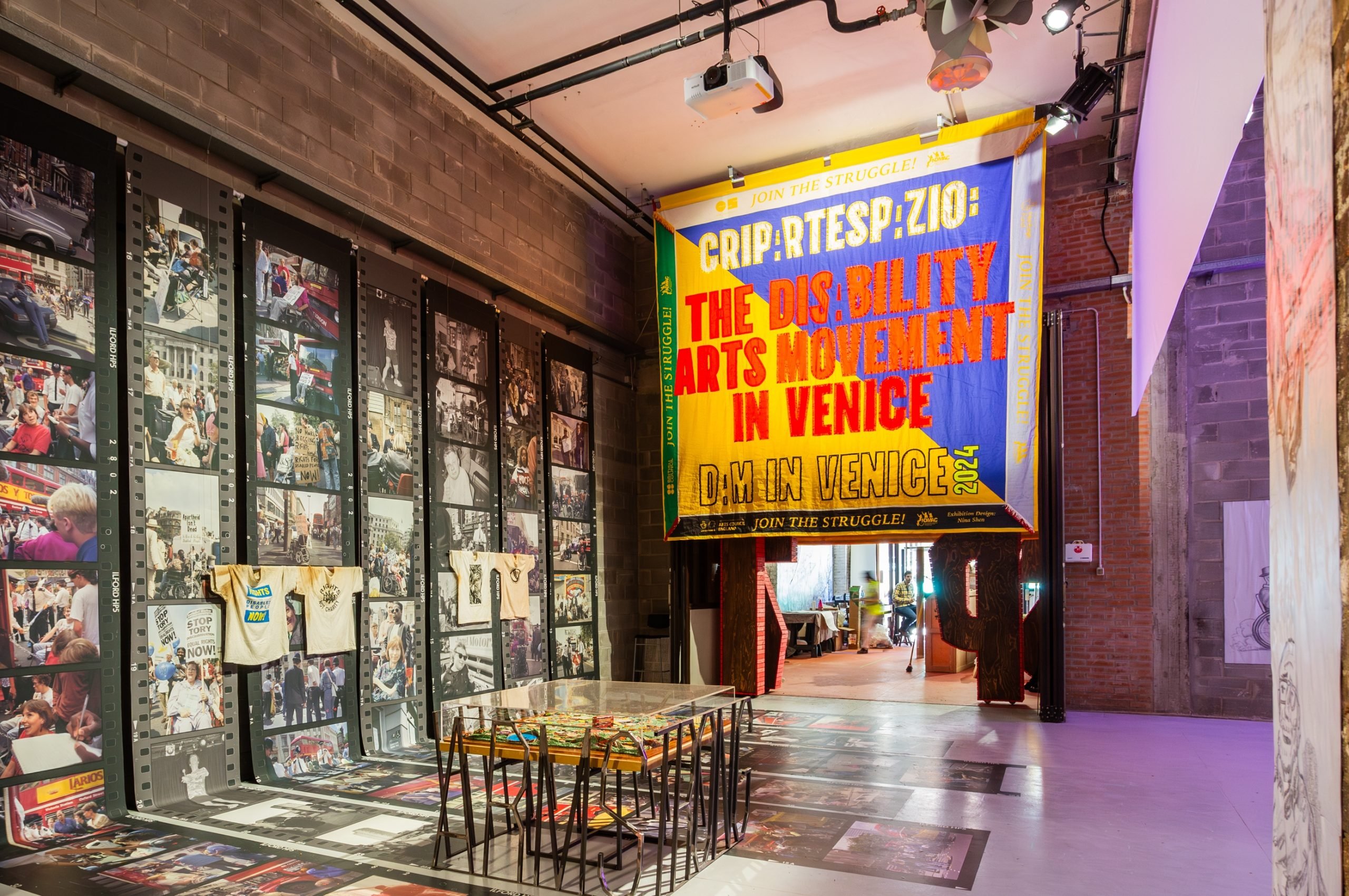 a large interior space with a chair and tables in it and some photos on the wall and a large textile with written "the disability arts movement in Venice"