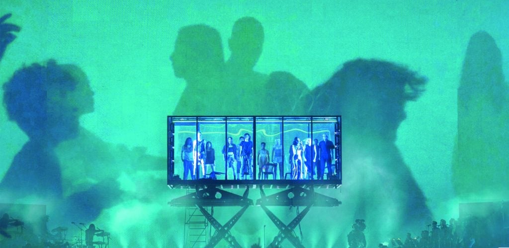 a large stage tinged in green and blue with a large rectangular transparent tank in the middle containing lots of figures silhouetted in blue
