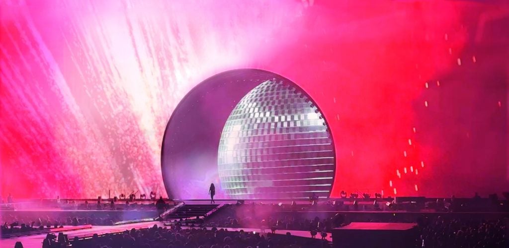 a huge pinky red stage with a circular cavity behind which we can see a disco ball, a womans figure in silhouette can also be seen on the stage