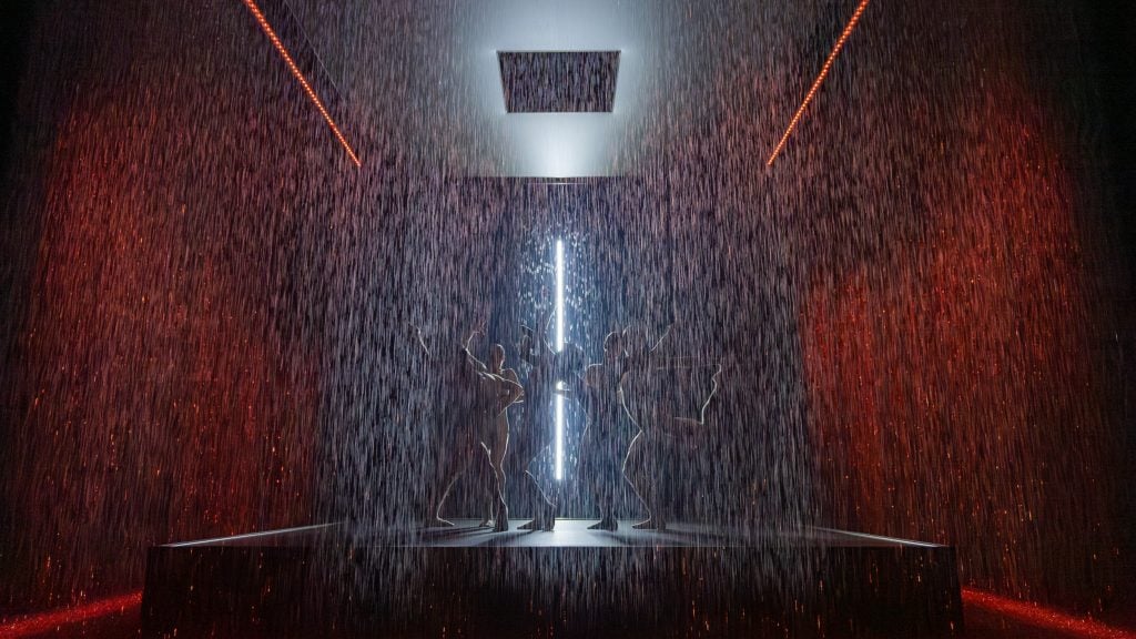 an interior space that appears to have rain falling and dancers can be seen behind the curtain of rain