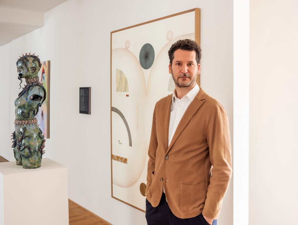 a man in a tan suit jacket stands next to a sculpture and in front of an abstract painting