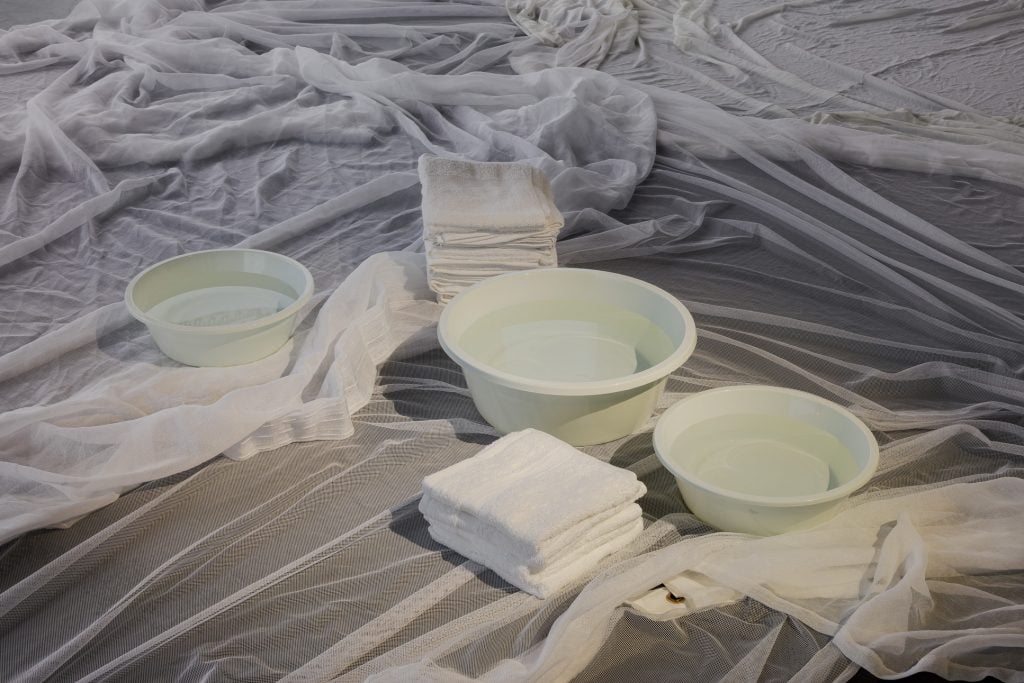 an installation view of an artwork included at liste art fair basel featuring a collection of bowls and two small piles of white wash clothes on top of white netting strewn on the ground.