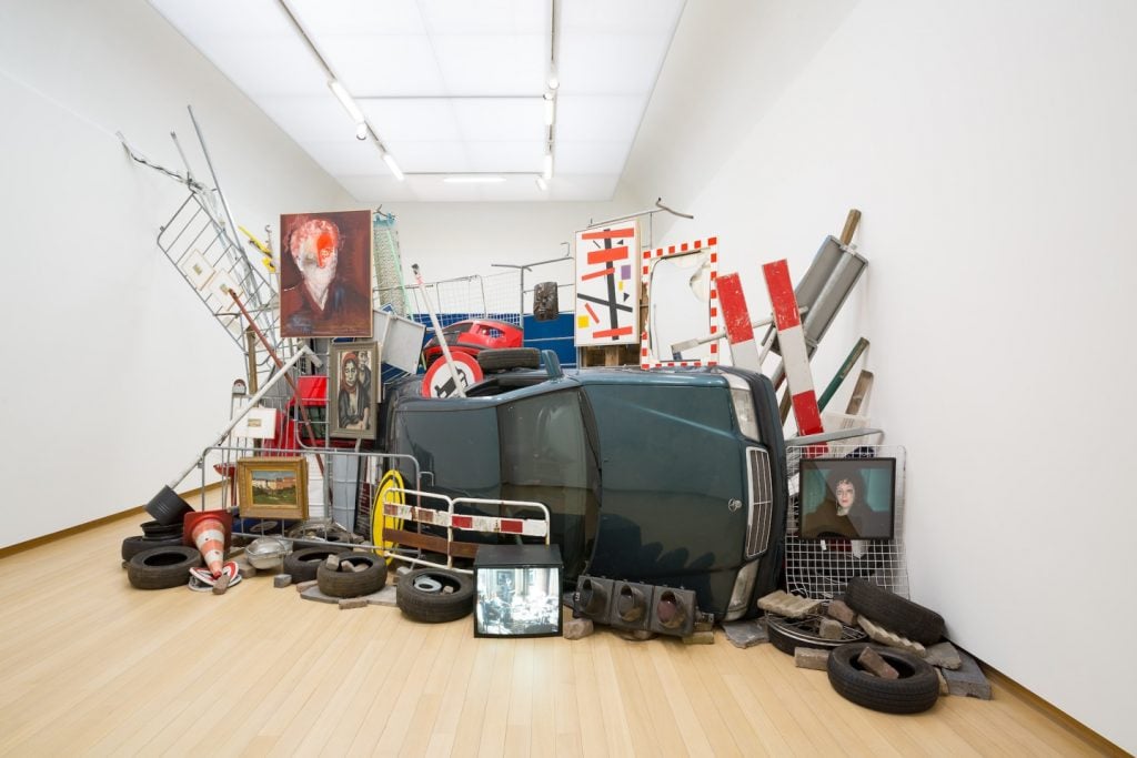 A modern art installation featuring an eclectic mix of objects including an overturned car, traffic cones, tires, metal barriers, and various signs. The arrangement includes several framed artworks, one of which depicts a person with an obscured face, and a television screen displaying an image of a person. The items are stacked and arranged in a seemingly chaotic manner against a white gallery wall with a wooden floor.
