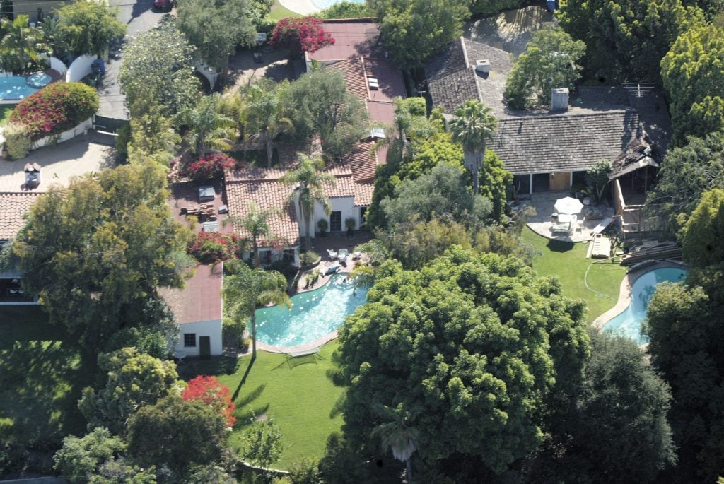 An aerial view of a lush residential area featuring multiple houses with tiled roofs, surrounded by dense greenery. The image showcases private swimming pools, well-maintained gardens, and a variety of trees and plants, creating a serene and secluded environment.