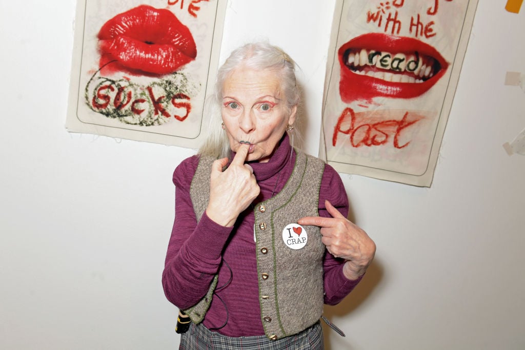 An older white woman places a finger in her mouth as she stands in front of a white wall.