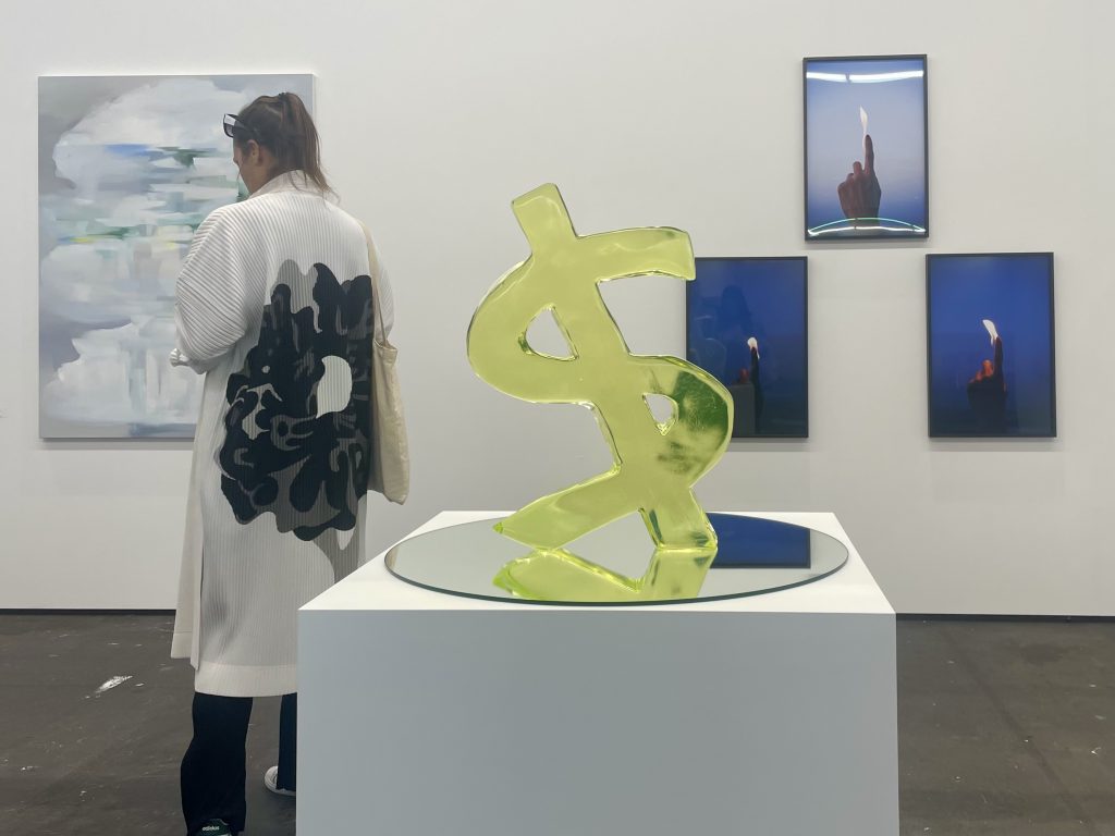 A sculpture in dollar sign at an art gallery booth at an art fair visited by a woman