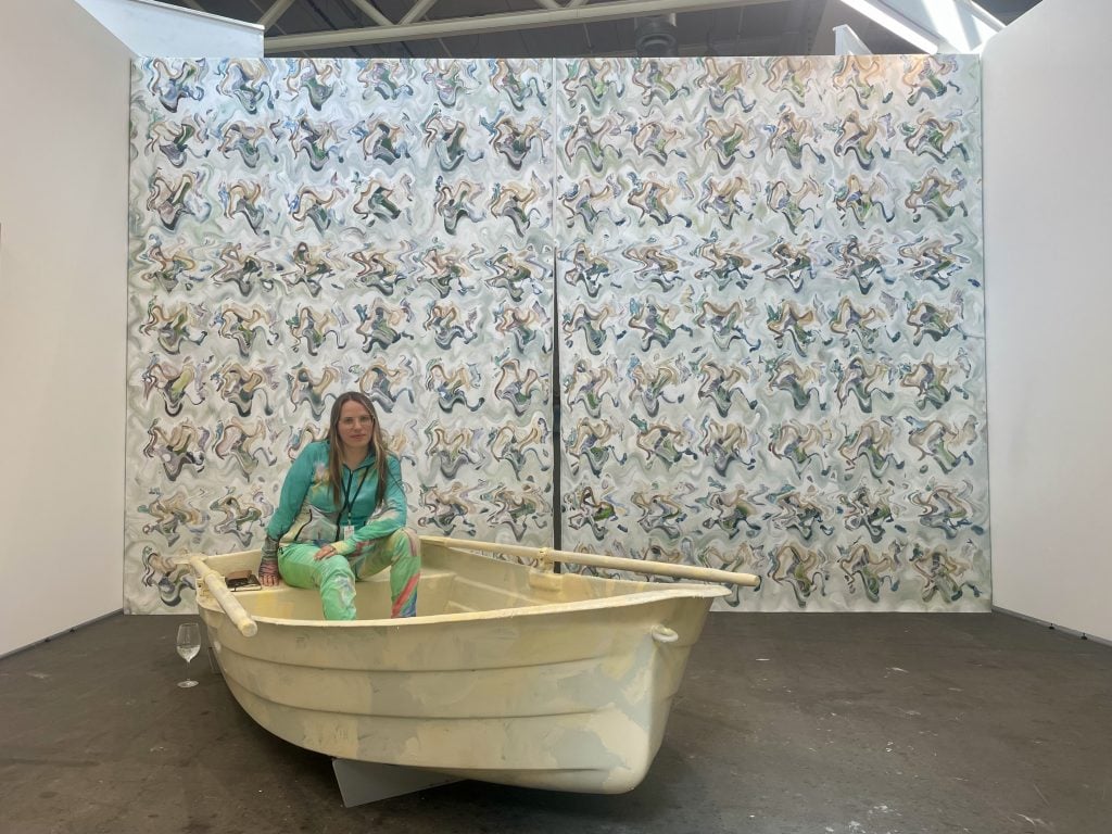 A woman in blonde long hair in a green track suit sitting on a boat against a backdrop of a large abstract painting