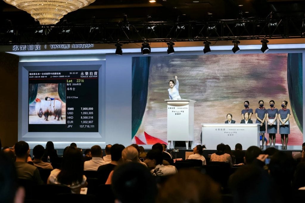 A color photo shows an auctioneer—a woman in a white outfit—soliciting bids in a large room.