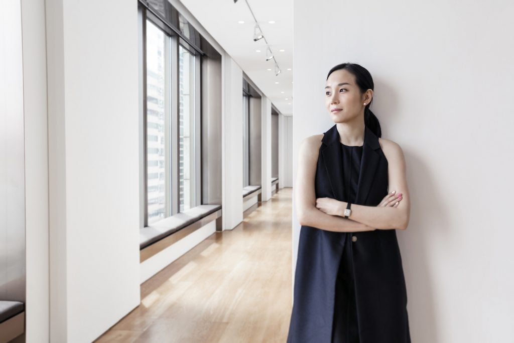 A woman in a long dark-colored dress leans against a white wall in a large, empty space.