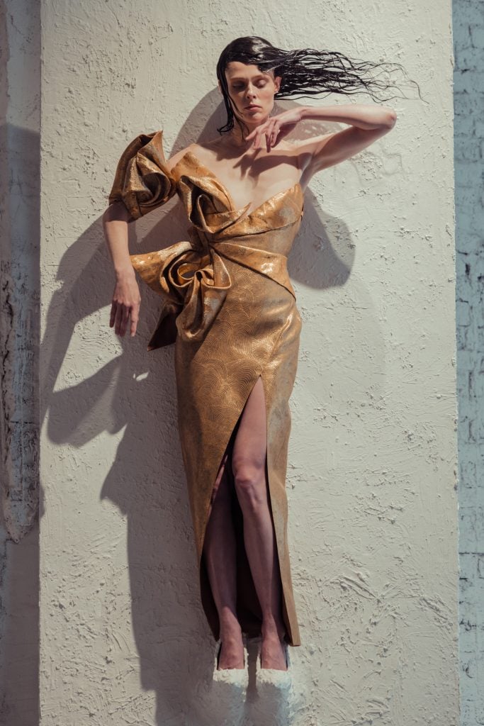 a woman is stuck to a white panel and is wearing a gold dress. her hair is stuck out to the side in an unnatural, stylised manner, she is doing a dramatic pose