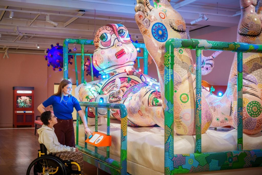 a large colorful sculpture of a human figure covered in doodles lying on a green hospital bed, its larger than life as wecan see because there are two people looking at it, one of whom is in a wheelchair