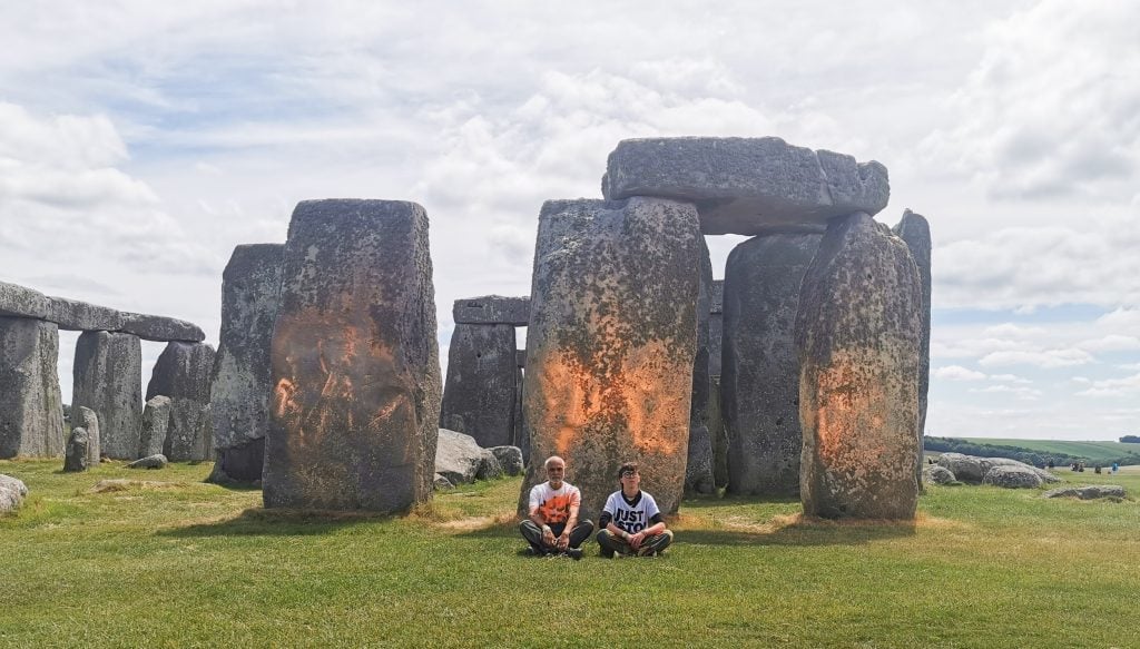 two people sit in front of a vast monolithic stone circle that has been sprayed with orange paint