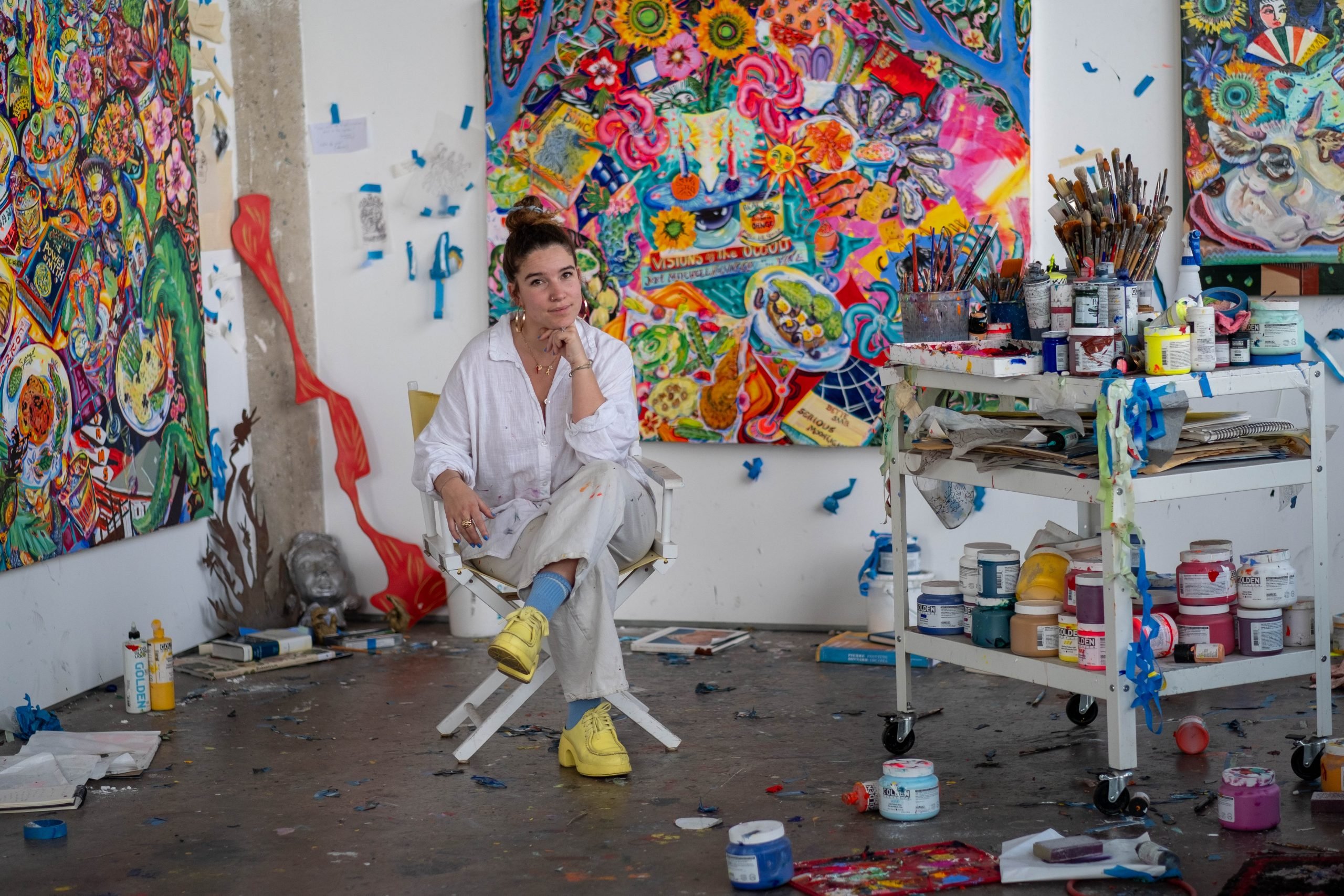 a white woman sits on a folding chair in front of a vibrantly colored painting