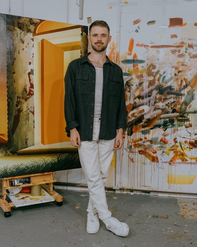 a white man in sneakers, white jeans, and a workman's shirt stands in front of a painting in progress and a studio wall covered in paint marks.