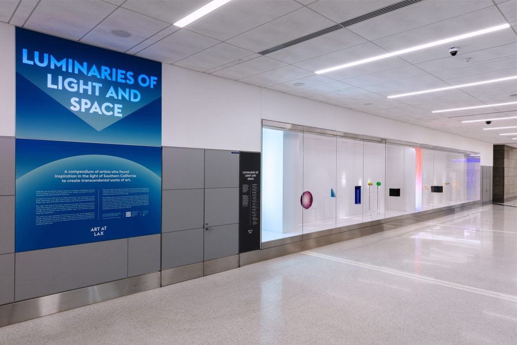 The image shows an art exhibit titled "Luminaries of Light and Space" at Los Angeles International Airport (LAX). The exhibit is dedicated to artists inspired by the unique light of Southern California, featuring a collection of contemporary and abstract artworks displayed in a long, glass-enclosed case. The exhibit aims to enhance the cultural experience for travelers, with a prominent blue sign providing an introduction and background information about the artists and their works. The installation includes various shapes and light-based pieces, creating a visually engaging corridor for airport visitors. The area is well-lit and designed to draw the attention of passersby.